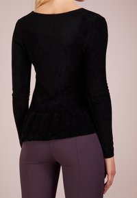 Zwarte lange mouw top van zacht, gestructureerd materiaal met een peplum zoom, gedragen over fitted donkerpaarse broeken. Glad ontwerp aan de achterkant, ronde hals.