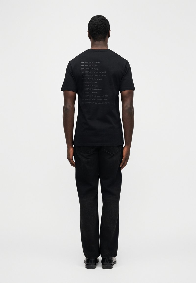 T-shirt en coton noir à encolure ronde, avec un texte blanc imprimé au dos. Design simple, porté avec un jean noir et des chaussures noires.