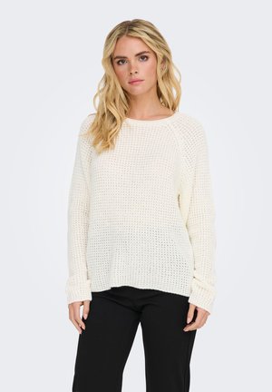Femme blonde portant un pull en maille blanc ample et un pantalon noir, debout devant un fond clair uni.