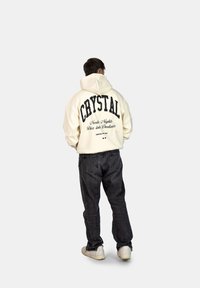 Felpa crema con la scritta "CRYSTAL" stampata in nero, con ulteriore testo. Indossata con jeans scuri e sneakers bianche, vista da dietro.