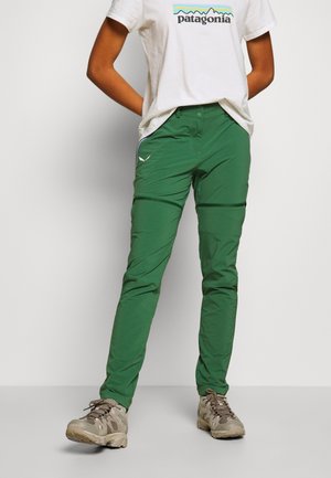 Osoba ubrana w zielone spodnie turystyczne z zamkami błyskawicznymi na kolanach, beżowe buty trekkingowe i białą koszulkę z logo Patagonia, stojąca z rękami za plecami.