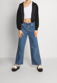 Hoodie nero a maniche corte sovrapposto a un top bianco a spalla scoperta, abbinato a jeans a gamba larga blu chiaro e sandali neri con piattaforma e fasce bianche.