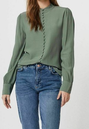 Femme portant un chemisier vert olive à manches longues avec des boutons recouverts de tissu et un jean bleu taille haute sur un fond uni.