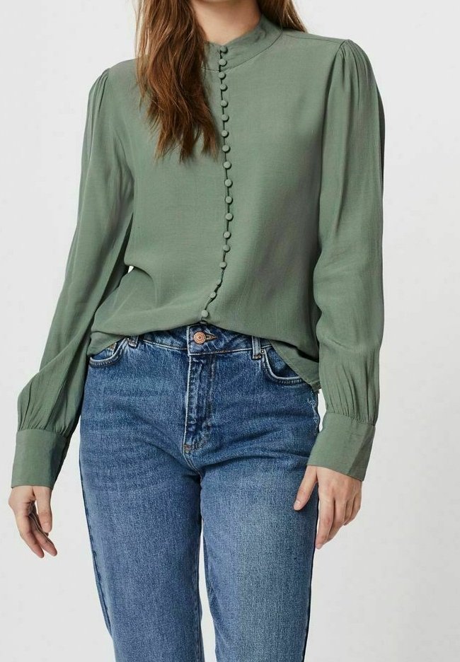 Femme portant un chemisier vert olive à manches longues avec des boutons recouverts de tissu et un jean bleu taille haute sur un fond uni.
