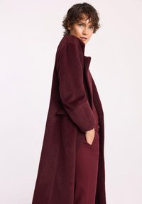 Bordowy płaszcz z mieszanki wełny o luźnym kroju, z wykładanym kołnierzem i bocznymi kieszeniami, noszony na dopasowany strój. Miękka tekstura, pełnej długości.