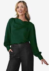 Blusa verde drappeggiata con maniche lunghe a sbuffo, texture liscia e scollatura ampia, abbinata a pantaloni in pelle nera a vita alta.