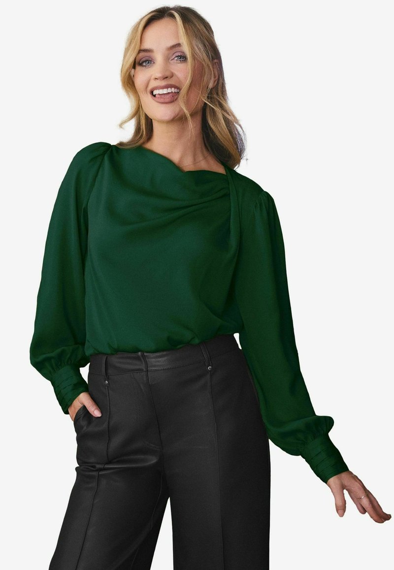 Blusa verde drappeggiata con maniche lunghe a sbuffo, texture liscia e scollatura ampia, abbinata a pantaloni in pelle nera a vita alta.