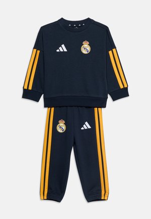 Survêtement bleu marine avec des bandes latérales jaunes, logo du Real Madrid sur la poitrine et la cuisse, et logos blancs Adidas sur le sweat-shirt et le pantalon.