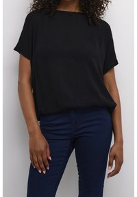 Sort kortærmet top med løs pasform og ribbet tekstur, parret med mørkeblå stramme jeans. Hænderne hviler på taljen.