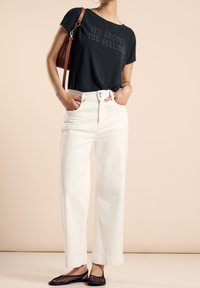 Uhlově šedé tričko s textem, bílé vysoké široké kalhoty, a otevřené černé baleríny z jemné síťoviny. Hnědá kabelka přes rameno, minimalistický design.