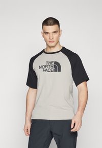 Camiseta de manga corta tipo raglán en gris con mangas negras, que presenta un destacado logo negro de "The North Face" en el frente.