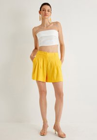 Witte strapless top en gele linnen hoge taille shorts met plooien, gecombineerd met beige bandjeshakken. De stof lijkt textuur te hebben en is lichtgewicht.