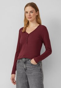 Rippiertes langärmeliges Shirt in Burgunderrot mit V-Ausschnitt und Knopfleiste. Kombiniert mit hochgeschnittenen grauen Jeans, was einen lässigen Look ergibt.