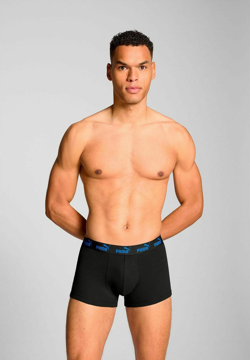 Black boxer briefs with a blue logo waistband, smooth fabric, fitted design, and a mid-rise style. Model stands with hands behind back.