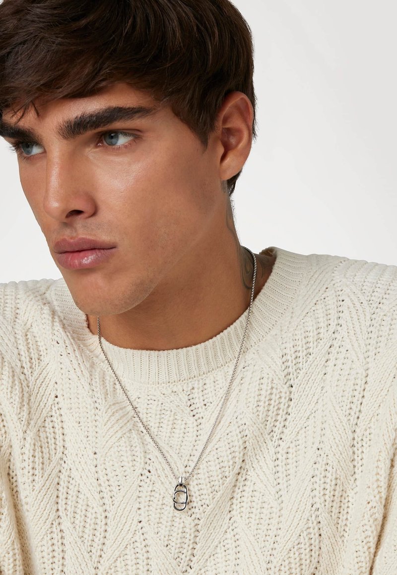 Collier en argent avec un pendentif ovale texturé, porté sur un pull crème en tricot à torsades. Le pendentif pend sur une chaîne fine.