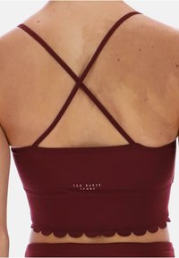 Bordowy sportowy crop top z falbaniastym wykończeniem, cienkimi krzyżującymi się ramiączkami i logo na plecach, wykonany z elastycznego materiału.
