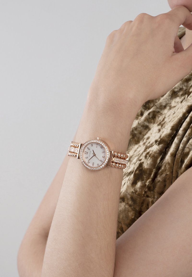 Polso di donna che indossa un orologio Guess in oro rosa con dettagli in cristallo e quadrante bianco, appoggiato vicino al mento su un indumento con trama in velluto.