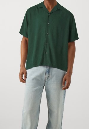 Mand iført en mørkegrøn kortærmet skjorte med knapper og lyseblå jeans, stående mod en ensfarvet lys baggrund.