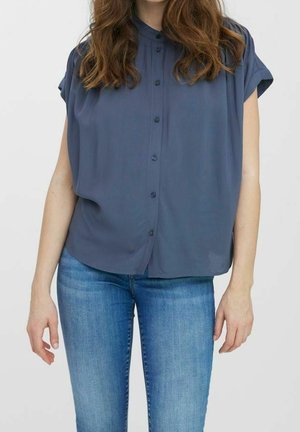 Femme portant un chemisier bleu foncé à manches courtes et à boutons, ainsi qu’un jean en denim bleu clair, debout devant un fond uni.