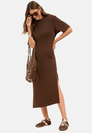 Kobieta w brązowej sukience midi, okularach przeciwsłonecznych, plecionej torbie i brązowych tenisówkach, stoi z jedną ręką na biodrze i uśmiecha się.