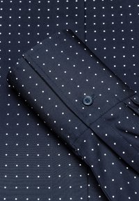 Navyblaue Bluse mit weißen Punkten, mit einem Knopf am Bündchen, glatte Textur und einem hochgeklappten Ärmel, der über dem gemusterten Stoff liegt.