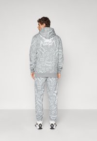 Mand iført grå Puma træningsdragt med hvid zigzag-mønster og Puma-logo på ryggen, vendende væk mod en ensfarvet lys baggrund.