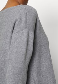 Grå stickad tröja med en bred, avskuren halsringning. Har ribbat mönster vid ärmkanterna och axlarna, vilket framhäver en avslappnad passform.