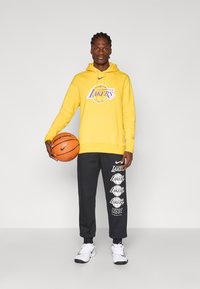 Fleece amarelo com impressão "Los Angeles Lakers", combinado com calças de moletom escuras com múltiplos logótipos dos Lakers. Modelo segura uma bola de basquete.