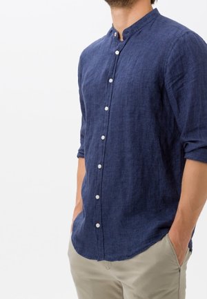 Mężczyzna w granatowej koszuli z długim rękawem na guziki z kołnierzykiem mandarynkowym i beżowych spodniach, ręce w kieszeniach.