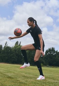 Svart treningstrøye, svarte shorts og lange svarte sokker. Iført hvite og røde Puma fotballsko. Dribler en rød og svart fotball.