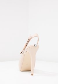 Scarpa beige con tacco alto, punta arrotondata, cinturino alla caviglia regolabile e un fiocco decorativo sul lato. Texture in pelle liscia con tacco lucido.