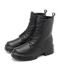 Une paire de bottines noires en cuir à lacets, avec de épais semelles en caoutchouc et des tirettes brunes, sur un fond blanc.