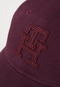 Gorra granate con tejido texturizado, visera ligeramente curvada y un logotipo geométrico bordado en hilo a juego en la parte frontal.