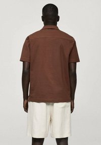 Polo marrón de manga corta, material de algodón, corte recto, con cuello y costura en el canesú trasero, combinado con pantalones cortos de lino de color claro.