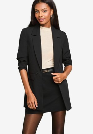 Černý blazer s přizpůsobeným designem, předním zapínáním na knoflík a jednou kapsou. Stylizovaný přes krémový top, spárovaný s černou sukní a opaskem.