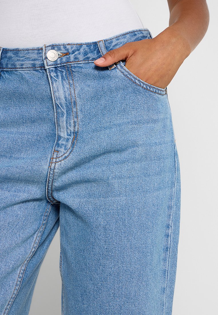 Jasnoniebieskie dżinsy z wysokim stanem, z przednią kieszenią i widocznym przeszyciem. Materiał ma gładkie, lekko teksturowane wykończenie.