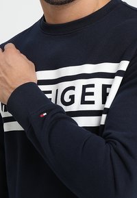 Muž v tmavě modré mikině s bílým horizontálním pruhem a výrazným černým textem, drží levou ruku na levém rameni.