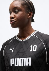Νεαρός Μαύρος με πλεγμένα μαλλιά, που φοράει μαύρη αθλητική φανέλα Puma με τον αριθμό 10, κοιτάζει προς τα πλάγια σε λευκό φόντο.