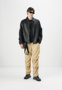Jachetă neagră din piele, cu fermoar, peste un tricou negru, asortată cu pantaloni cargo bej, cu buzunare, și adidași negri. Accesoriile includ ochelari de soare.