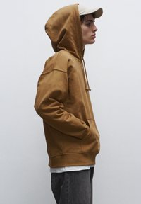 Felpa marrone con cappuccio e cordino, tasca a marsupio e polsini a coste, indossata sopra una t-shirt bianca, abbinata a jeans grigi e un cappello beige.