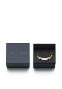Bracciale in metallo rosa con un design liscio e curvo, posizionato all'interno di una scatola blu navy con interno nero e testo dorato.
