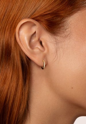 Pendiente de aro dorado con un acabado suave y pulido, llevado en el lóbulo de la oreja, acentuado contra el cabello rojo y un tono de piel claro.