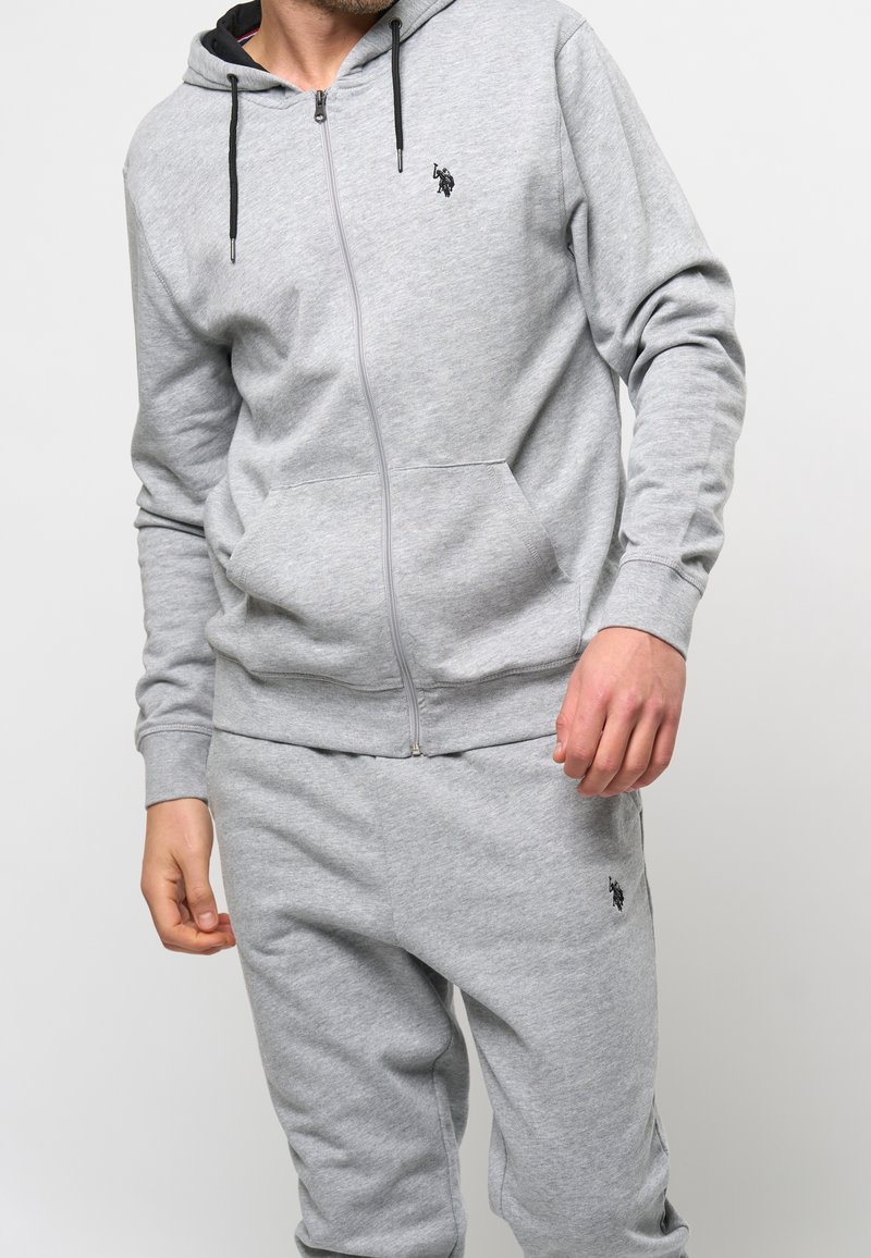 Harmaa vetoketjullinen huppari ja vastaavat collegehousut pehmeästä, teksturoidusta kankaasta. Hupparissa on etutasku ja mustat kiristysnyörit. Rinnassa pieni logo.