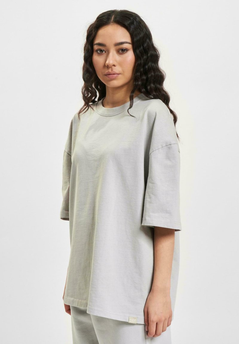 Grey oversized t-shirt with short sleeves, round neckline, and subtle texture. Features a small logo tag on the lower left side.