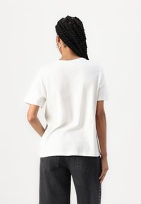 T-shirt en coton blanc avec un col rond et des manches courtes, vu de dos. Associé à un jean foncé avec des détails de surpiqûres subtils.