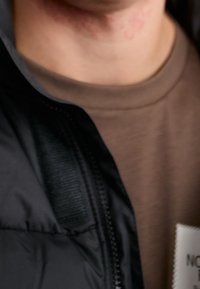 Schwarze gepolsterte Jacke mit einem Reißverschluss, die aus strukturiertem Stoff besteht und darunter ein glattes, rundes braunes T-Shirt trägt. Die Etiketten sind auf der Jacke sichtbar.