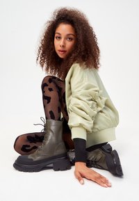 Bottes de combat vert olive à lacets avec une semelle noire épaisse, assorties à des leggings noirs transparents à imprimé léopard. Modèle assis, portant une veste bomber vert clair.