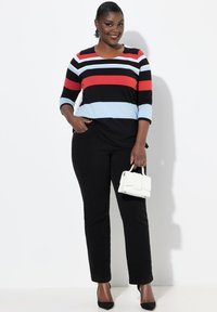 Top de manga larga a rayas blanco y negro con rayas rojas y azul claro, combinado con pantalones negros. La modelo sostiene un pequeño bolso blanco.