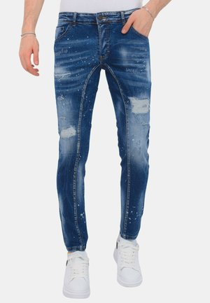 Slim-fit blauwe distressed jeans met verfspatten, gedragen met witte sneakers en een wit top, handen gedeeltelijk zichtbaar.