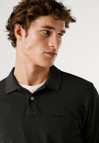 Černé polo tričko vyrobené z texturované tkaniny s límečkem a malým vyšitým logem. Obsahuje zapínání na knoflíky a vrstvené bílé tričko pod ním.